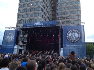 A stage surrounded by blue signs with a dog in a suit and the words Boston Calling. A band is on the stage and a large crowd is in front of them.